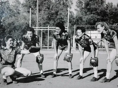 A group photo of women in softball uniforms wearing baseball gloves pose for a photo