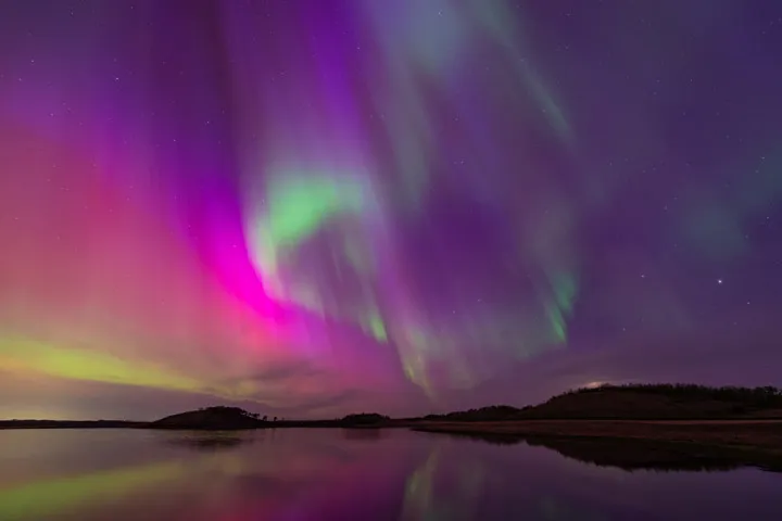 purple and green streaks in the air over water and mountains at night