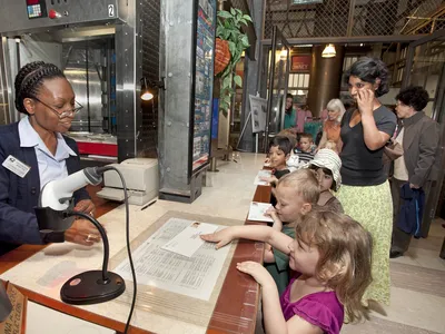 A group of students wait in line at a Post Office. Each student has a stamped envelope they are handing to the postal worker behind the counter.